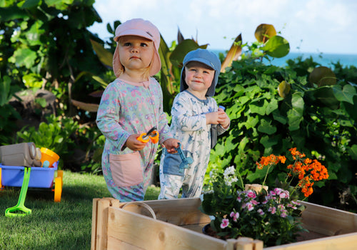 Two babies playing outdoors in a Hawaiian backyard, wearing UV Skinz sun flap hats for sun protection, in front of a vibrant planter box.