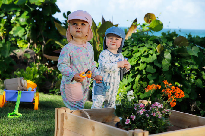 Two babies playing outdoors in a Hawaiian backyard, wearing UV Skinz sun flap hats for sun protection, in front of a vibrant planter box.