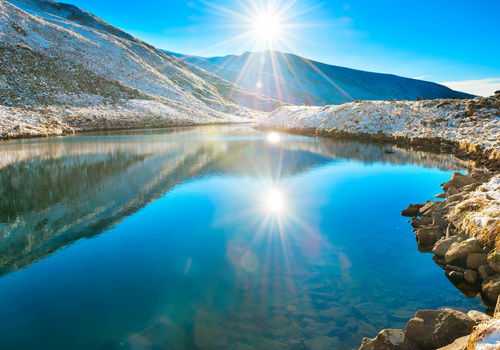 Sunrise over a serene winter lake with snow-capped mountains, highlighting the importance of UV protection; a person wearing UV Skinz UPF clothing stands at the lakeshore, emphasizing sun safety in winter activities.