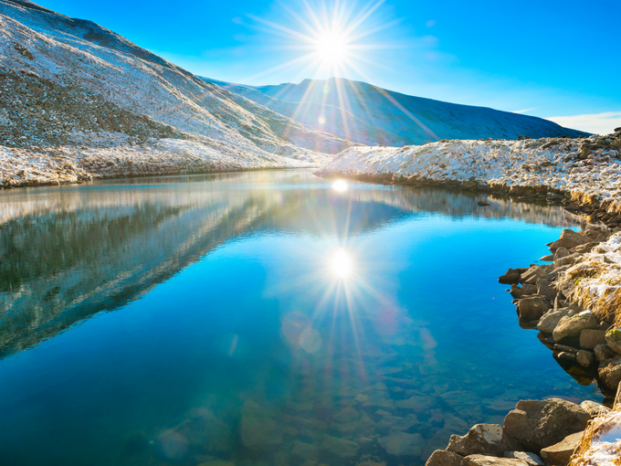 Sunrise over a serene winter lake with snow-capped mountains, highlighting the importance of UV protection; a person wearing UV Skinz UPF clothing stands at the lakeshore, emphasizing sun safety in winter activities.