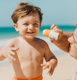 Family sun safety in action: A loving mother wearing UV Skinz beachwear protects her infant son from potential sun damage by carefully applying protective sunscreen during their beach day